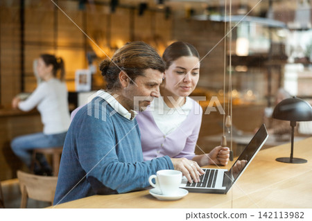 Portrait of middle-aged male and young female surfing Internet on notebook while drinking tea or coffee in cafeteria Portrait of middle-aged male and young female surfing Internet on notebook while drinking tea or coffee in cafeteria 142113982