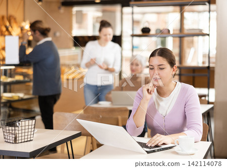 Positive European young woman drinking and enjoying coffee while working on laptop in cafe. Freelance and remote work Positive European young woman drinking and enjoying coffee while working on laptop in cafe. Freelance and remote work 142114078