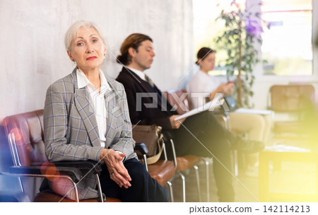 Serious-minded female candidate siting on chair and waiting for job interview in hallway Serious-minded female candidate siting on chair and waiting for job interview in hallway 142114213