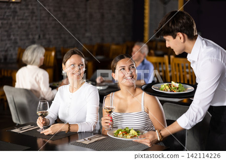 Young waiter serving dishes to female guests in restaurant Young waiter serving dishes to female guests in restaurant 142114226