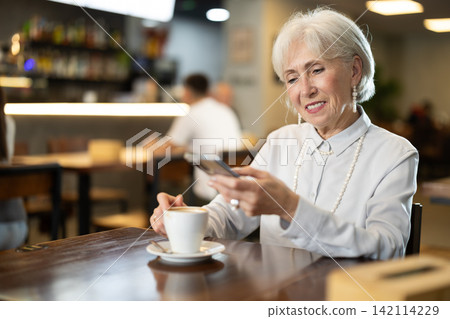 Elderly woman with smartphone drinking coffee at table in cafe Elderly woman with smartphone drinking coffee at table in cafe 142114229