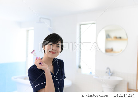 一位女士在浴室裡面帶著微笑,手裡拿著刮鬍刀。 一位女士在浴室裡面帶著微笑,手裡拿著刮鬍刀。 142114234