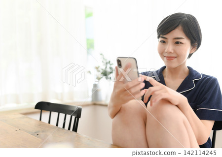 一名身穿睡衣的女子一邊操作智慧型手機一邊微笑。 一名身穿睡衣的女子一邊操作智慧型手機一邊微笑。 142114245