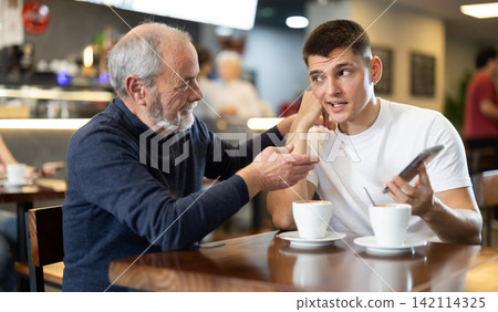 Elderly man comforts an upset guy while sitting at table in cafe Elderly man comforts an upset guy while sitting at table in cafe 142114325