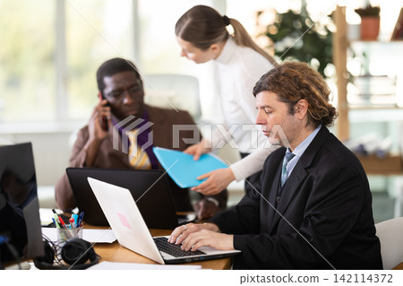 Woman with documents is standing next to African male colleague with laptop Woman with documents is standing next to African male colleague with laptop 142114372