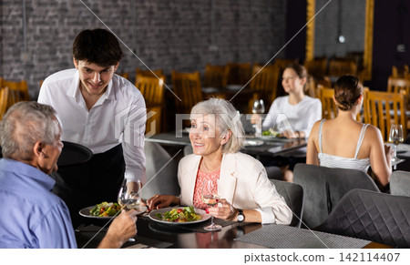 Young waiter serving dishes to aged man and woman in restaurant Young waiter serving dishes to aged man and woman in restaurant 142114407