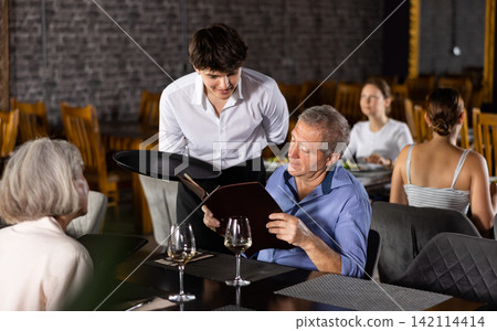 Young waiter recommending dishes from menu to elderly man Young waiter recommending dishes from menu to elderly man 142114414
