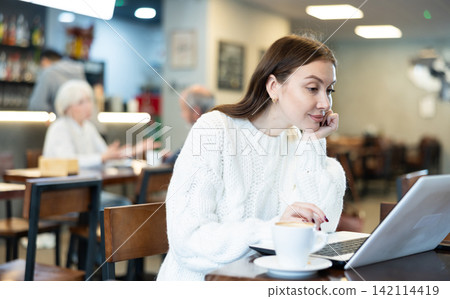 In cozy cafe hall, freelance woman using laptop and drinks coffee. In cozy cafe hall, freelance woman using laptop and drinks coffee. 142114419
