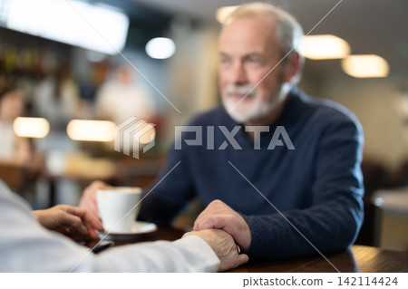 Elderly man sitting holding elderly womans hand in coffee shop Elderly man sitting holding elderly womans hand in coffee shop 142114424