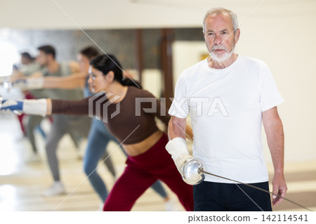 Portrait of an elderly man holding rapier during fencing training in gym Portrait of an elderly man holding rapier during fencing training in gym 142114541