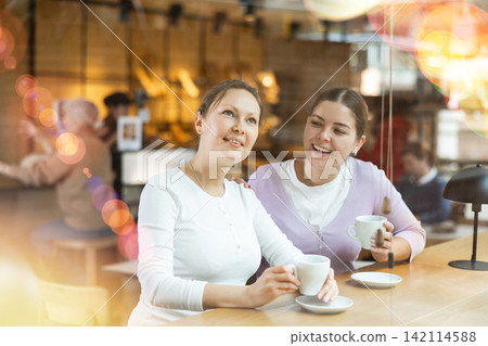 Two cheerful female friends enjoying their coffee in cozy cafe Two cheerful female friends enjoying their coffee in cozy cafe 142114588