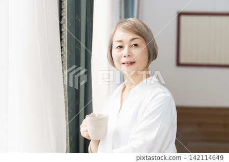 一位老婦人在客廳窗邊喝咖啡。 142114649