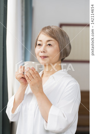 A senior woman drinking coffee by the living room window. A senior woman drinking coffee by the living room window. 142114654