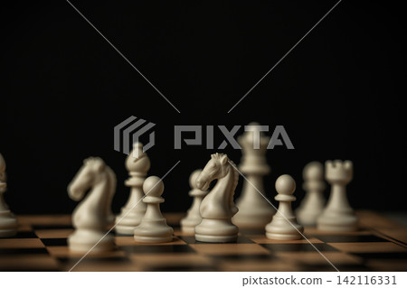 white chess knight standing on a board, surrounded by blurred pieces on black background white chess knight standing on a board, surrounded by blurred pieces on black background 142116331