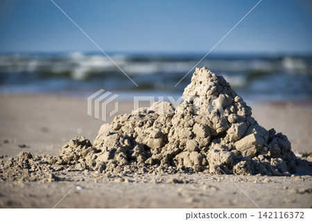 Sandcastle Mound on Sandy Beach Shore with Blurred Ocean Waves and Blue Sky Background Sandcastle Mound on Sandy Beach Shore with Blurred Ocean Waves and Blue Sky Background 142116372