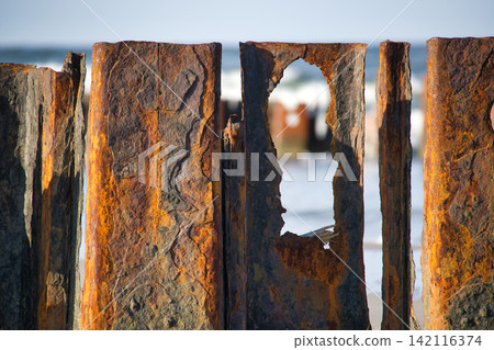 Rusty Corrugated Metal Sheet with Holes Standing on Beach Against Ocean Waves and Blue Sky Rusty Corrugated Metal Sheet with Holes Standing on Beach Against Ocean Waves and Blue Sky 142116374