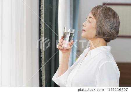 A senior woman drinking water from a glass by the living room window. 142116707