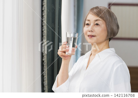 A senior woman drinking water from a glass by the living room window. A senior woman drinking water from a glass by the living room window. 142116708