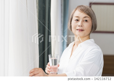 A senior woman drinking water from a glass by the living room window. A senior woman drinking water from a glass by the living room window. 142116712