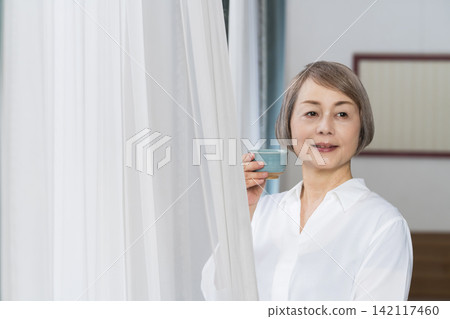 一位老婦人坐在客廳窗邊,用茶杯喝茶。 一位老婦人坐在客廳窗邊,用茶杯喝茶。 142117460