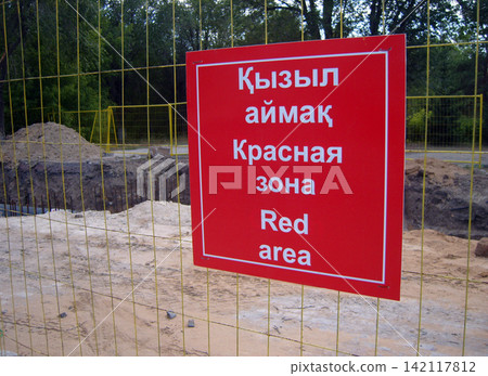 A red warning sign is posted on a yellow mesh fence surrounding a deep construction excavation. The sign text, in three languages, reads: Red area. Trees stand in the background. A red warning sign is posted on a yellow mesh fence surrounding a deep construction excavation. The sign text, in three languages, reads: Red area. Trees stand in the background. 142117812