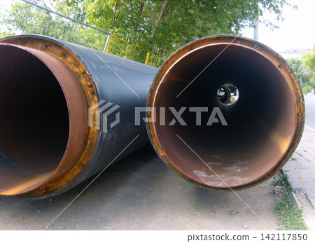 Large, industrial-grade insulated pipes lie horizontally at roadside construction site. Inner metal surfaces, distinctly rusty, with yellow foam insulation separating them from the dark outer casing Large, industrial-grade insulated pipes lie horizontally at roadside construction site. Inner metal surfaces, distinctly rusty, with yellow foam insulation separating them from the dark outer casing 142117850