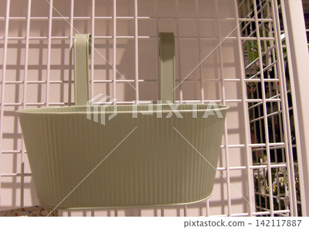 A close-up view of a pale, light-green ribbed metal planter pot hanging from a white wire mesh panel with two hooks. The corrugated container is displayed in an indoor setting with grid walls. A close-up view of a pale, light-green ribbed metal planter pot hanging from a white wire mesh panel with two hooks. The corrugated container is displayed in an indoor setting with grid walls. 142117887