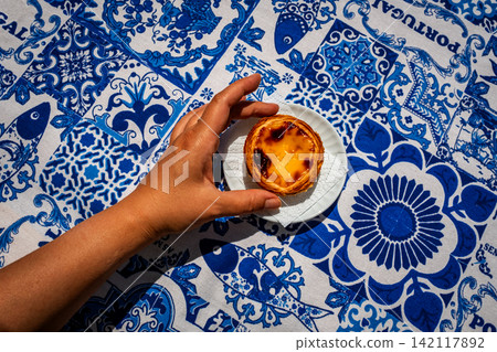 Top down food shot of tanned female hand placing one pastel de nata custard tart on white saucer on blue and white tablecloth with traditional Portuguese azulejo tile motifs with simple copy space 142117892
