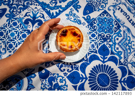 One pastel de nata custard tart on white saucer served by tanned female hand on blue and white tablecloth with traditional Portuguese azulejo tile motifs on sunny day in high key. Regional delicacy 142117893