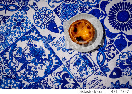 Flat-lay view of one pastel de nata custard tart on white plate on blue and white tablecloth with traditional Portuguese azulejo tile motifs with simple copy space. Translation: best things in life Flat-lay view of one pastel de nata custard tart on white plate on blue and white tablecloth with traditional Portuguese azulejo tile motifs with simple copy space. Translation: best things in life 142117897