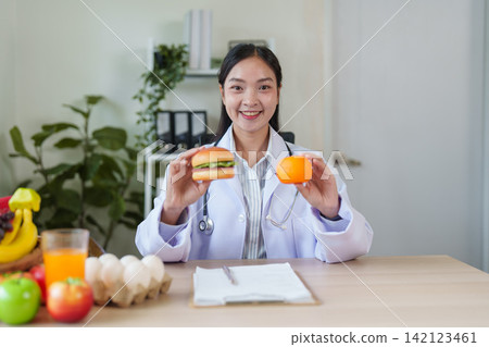 Asian nutritionist doctor holding hamburger and orange for healthy diet choice Asian nutritionist doctor holding hamburger and orange for healthy diet choice 142123461