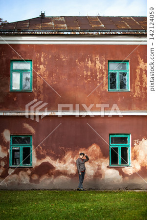 Young stylish person in a tweed jacket and flat cap standing near a large old red wall with green windows. Urban street fashion, vintage architecture facade, fall lifestyle, and autumn city lifestyle 142124059