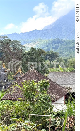 Traditional Balinese temple roofs and a rural house appear nestled among dense tropical vegetation. Looking out towards the majestic Mount Agung in the distance Traditional Balinese temple roofs and a rural house appear nestled among dense tropical vegetation. Looking out towards the majestic Mount Agung in the distance 142124110