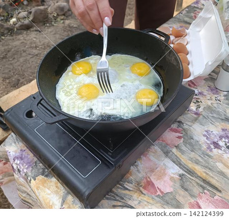 High angle close up of a hand with manicured pink nails stirring four frying eggs in a black cast iron pan on a portable stovetop, beside an open brown egg carton on a floral tablecloth outdoors... High angle close up of a hand with manicured pink nails stirring four frying eggs in a black cast iron pan on a portable stovetop, beside an open brown egg carton on a floral tablecloth outdoors... 142124399