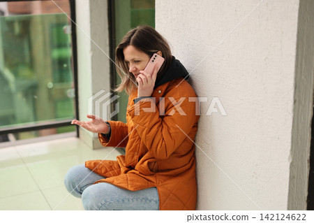 Woman in orange coat sitting beside wall and talking on smartphone while gesturing with open hand indoors near bright window, showing concern and waiting for response Woman in orange coat sitting beside wall and talking on smartphone while gesturing with open hand indoors near bright window, showing concern and waiting for response 142124622