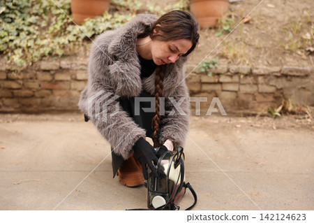 Young woman crouching outdoors while checking transparent backpack beside stone wall, wearing furry coat, long braid, gloves, and boots in soft natural light with autumn tones 142124623