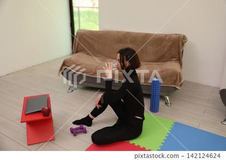 Woman in black workout clothing sits on colorful exercise mat at home, drinking water beside dumbbells, laptop, apple, foam roller, and sofa in bright room after fitness session Woman in black workout clothing sits on colorful exercise mat at home, drinking water beside dumbbells, laptop, apple, foam roller, and sofa in bright room after fitness session 142124624