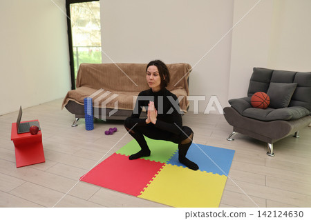 Woman practicing yoga squat pose on colorful exercise mat in living room, with laptop, fitness accessories, sofa, and basketball nearby, showing home workout routine, balance, focus, and wellness Woman practicing yoga squat pose on colorful exercise mat in living room, with laptop, fitness accessories, sofa, and basketball nearby, showing home workout routine, balance, focus, and wellness 142124630