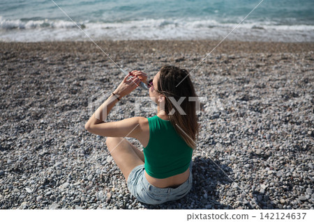 Woman sitting on pebble beach beside calm sea, using sun protection stick and enjoying bright summer sunlight with relaxed back view, coastal travel mood, natural outdoor leisure scene Woman sitting on pebble beach beside calm sea, using sun protection stick and enjoying bright summer sunlight with relaxed back view, coastal travel mood, natural outdoor leisure scene 142124637