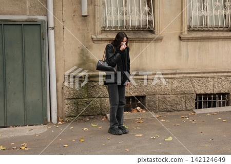 Woman in black coat standing beside weathered city wall, holding tissue to face near quiet sidewalk with autumn leaves, worn windows, old doorway, and muted urban textures Woman in black coat standing beside weathered city wall, holding tissue to face near quiet sidewalk with autumn leaves, worn windows, old doorway, and muted urban textures 142124649