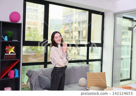 Smiling young woman standing indoors with hands clasped in bright modern apartment, framed by large windows, cozy sofa, shelves, gifts, and daylight creating relaxed home atmosphere Smiling young woman standing indoors with hands clasped in bright modern apartment, framed by large windows, cozy sofa, shelves, gifts, and daylight creating relaxed home atmosphere 142124653