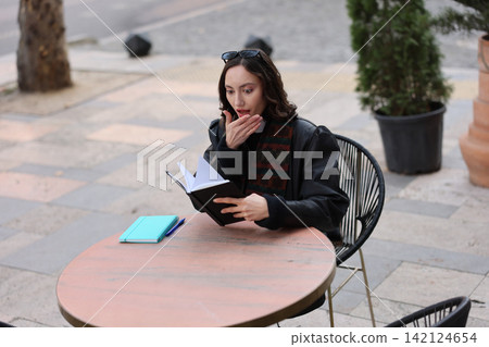 Thoughtful young woman reading notebook outdoors at caf table, looking surprised while holding hand near mouth, with pen and second notebook on table in soft daylight Thoughtful young woman reading notebook outdoors at caf table, looking surprised while holding hand near mouth, with pen and second notebook on table in soft daylight 142124654