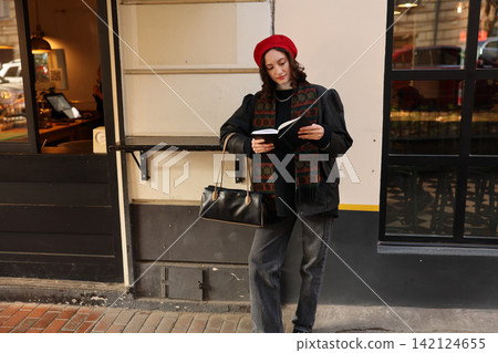 Young woman in red beret reading notebook beside cafe entrance, wearing black outfit, patterned scarf, and shoulder bag on quiet city sidewalk with warm storefront reflections Young woman in red beret reading notebook beside cafe entrance, wearing black outfit, patterned scarf, and shoulder bag on quiet city sidewalk with warm storefront reflections 142124655