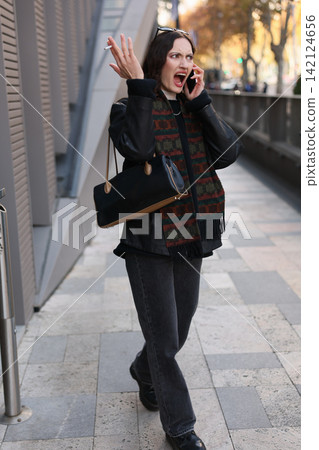 Frustrated woman shouting during phone call on city sidewalk, gesturing with cigarette, dark coat, patterned scarf, handbag, autumn street scene with strong urban tension Frustrated woman shouting during phone call on city sidewalk, gesturing with cigarette, dark coat, patterned scarf, handbag, autumn street scene with strong urban tension 142124656