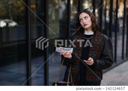 Young woman standing outdoors near glass storefront, holding cash and cards while looking upward with thoughtful expression, dressed in dark clothing with patterned scarf and handbag in city Young woman standing outdoors near glass storefront, holding cash and cards while looking upward with thoughtful expression, dressed in dark clothing with patterned scarf and handbag in city 142124657