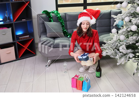 Young woman in Santa hat and red dress kneels beside decorated Christmas tree, holding stacked gift boxes in modern living room with festive ornaments, wrapping bags, bottle, and soft winter holiday 142124658