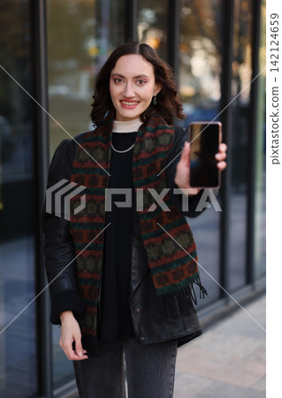 Smiling young woman taking selfie with smartphone outdoors beside glass building, wearing dark coat and patterned scarf, holding phone toward camera in urban setting with soft natural light Smiling young woman taking selfie with smartphone outdoors beside glass building, wearing dark coat and patterned scarf, holding phone toward camera in urban setting with soft natural light 142124659