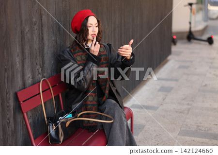 Young woman in red beret applying lipstick while sitting on outdoor bench beside dark wooden wall, carrying handbag and wearing layered winter outfit on city sidewalk Young woman in red beret applying lipstick while sitting on outdoor bench beside dark wooden wall, carrying handbag and wearing layered winter outfit on city sidewalk 142124660