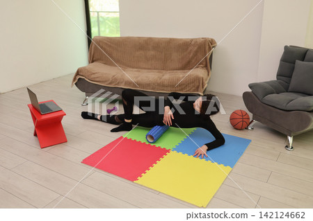 Woman performing floor stretch exercise on colorful mat in bright living room with laptop, foam roller, dumbbell, and basketball nearby, creating calm home fitness scene Woman performing floor stretch exercise on colorful mat in bright living room with laptop, foam roller, dumbbell, and basketball nearby, creating calm home fitness scene 142124662