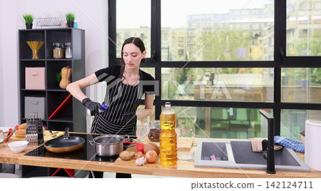 Young woman cooking in modern kitchen with apron and gloves, preparing meal at wooden counter with fresh ingredients, cookware, stove, city view window, natural daylight, home food preparation Young woman cooking in modern kitchen with apron and gloves, preparing meal at wooden counter with fresh ingredients, cookware, stove, city view window, natural daylight, home food preparation 142124711